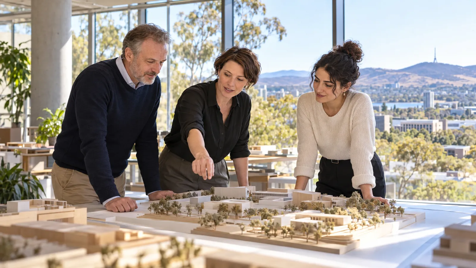 Zwei Frauen und ein Mann besprechen ein Architekturmodell in einem hellen Büro mit Blick auf Canberra.